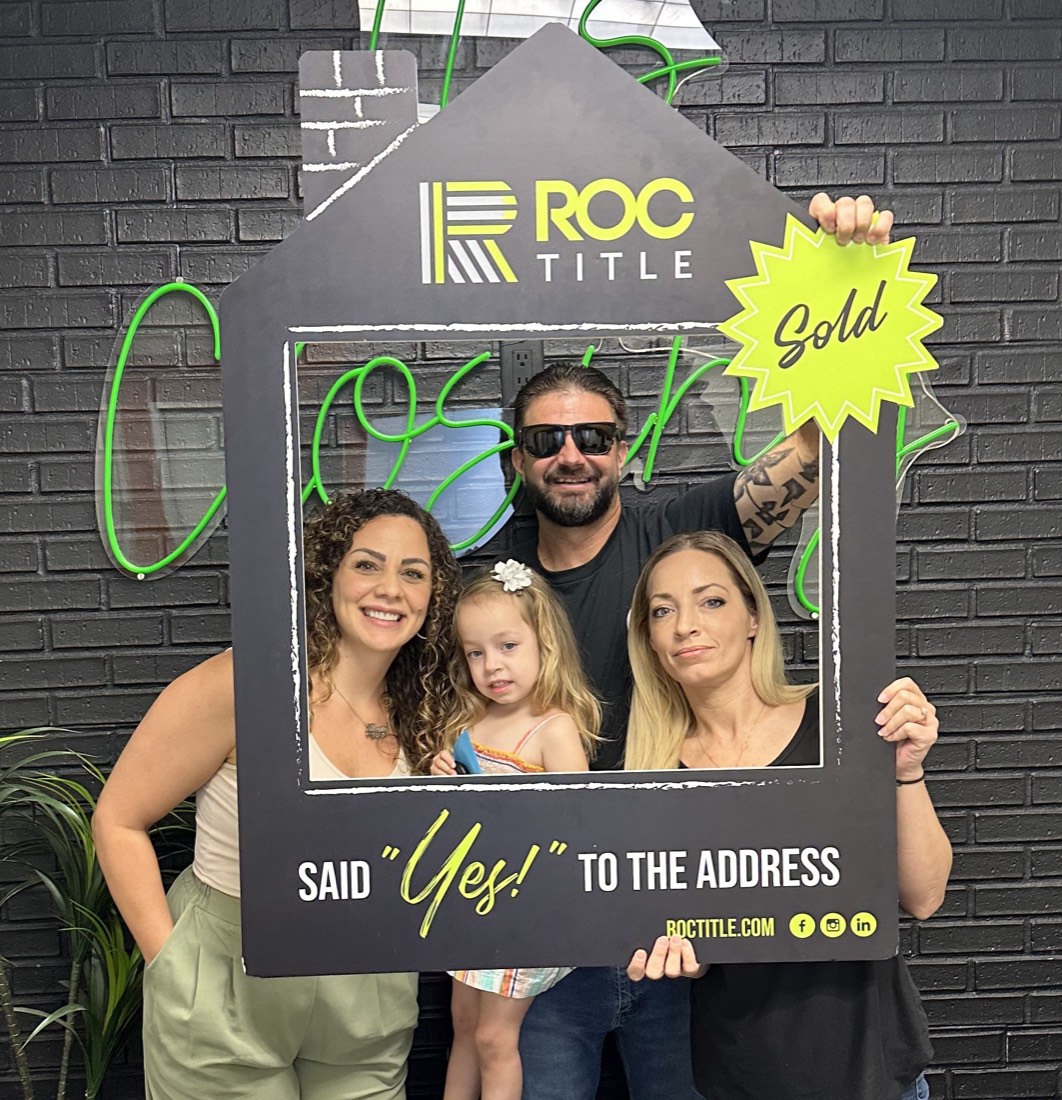 Alexis celebrating with a family holding a "Said Yes to the Address" sign on closing day